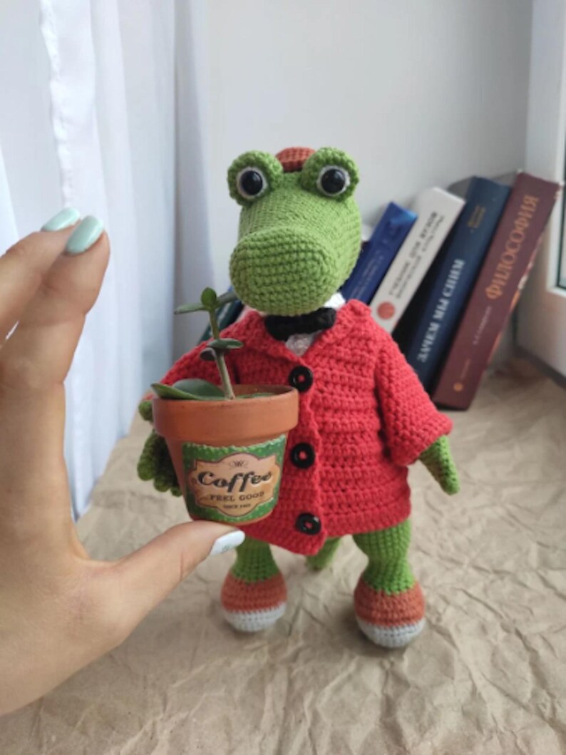 Puede incluir: Un cocodrilo de crochet verde que lleva un c&aacute;rdigan rojo y una pajarita negra sostiene una peque&ntilde;a maceta de terracota con una planta verde y una etiqueta que dice "Coffee Feel Good".