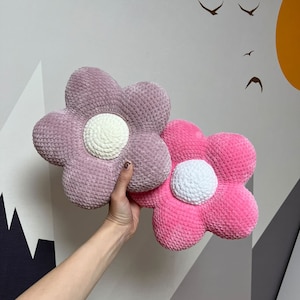 Könnte beinhalten: Zwei gehäkelte Kissen in Blumenform. Eines ist rosa und das andere ist helllila. Beide haben eine weiße gehäkelte Mitte.