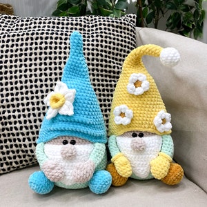 May include: Two handmade crochet gnomes. One gnome has a light blue hat and body with a white and yellow flower. The other gnome has a yellow hat and body with white flower accents. Both gnomes have white beards and tan noses.