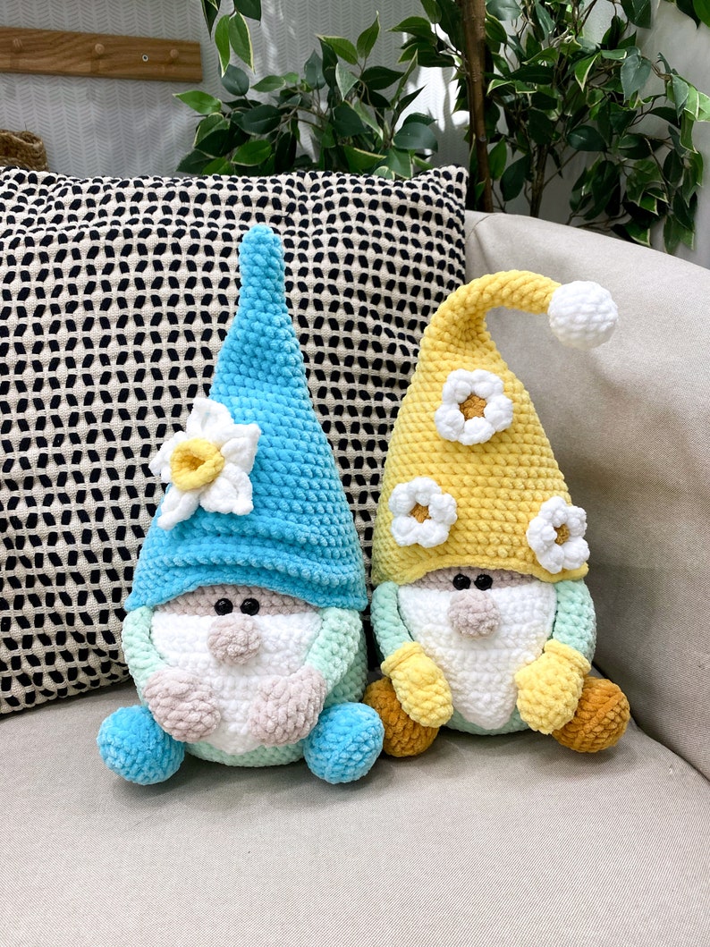 May include: Two handmade crochet gnomes. One gnome has a light blue hat and body with a white and yellow flower. The other gnome has a yellow hat and body with white flower accents. Both gnomes have white beards and tan noses.
