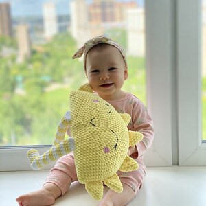 May include: A baby wearing a pink and white floral headband holds a yellow crocheted sun-shaped plush toy. The toy has a smiling face with closed eyes and a pink flower on its cheek.