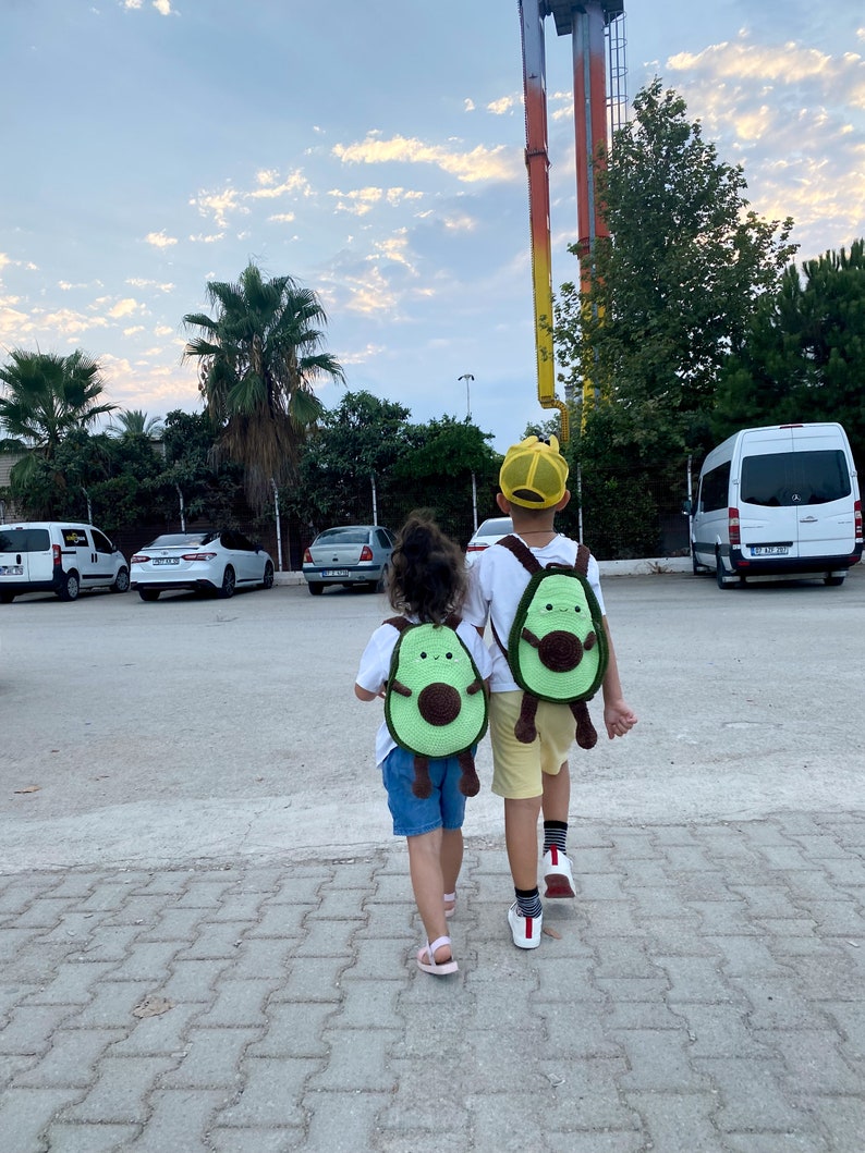 May include: Two children wearing avocado-shaped knitted backpacks walk away from the camera. The backpacks have brown seed details and smiling faces. The children are wearing casual clothing and are walking on a brick pathway.