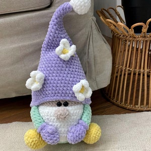 May include: A handmade gnome doll with a lavender hat decorated with white flower accents. The gnome has a white beard, a tan nose, and black eyes. Its body is a mix of pastel colours, including green, purple, and yellow.
