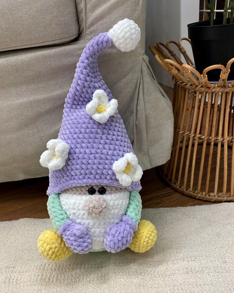 May include: A handmade gnome doll with a lavender hat decorated with white flower accents. The gnome has a white beard, a tan nose, and black eyes. Its body is a mix of pastel colours, including green, purple, and yellow.