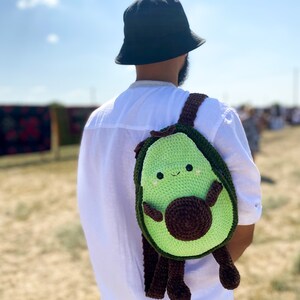 May include: A person wearing a black bucket hat and a white shirt is carrying a green and brown crocheted avocado backpack with a smiling face.