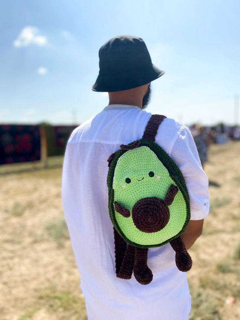 May include: A person wearing a black bucket hat and a white shirt is carrying a green and brown crocheted avocado backpack with a smiling face.