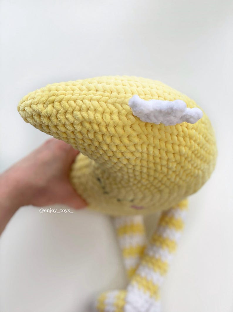 May include: A handmade yellow crochet moon plush toy with a white cloud accent. The moon has a smiling face and striped yellow and white legs. The toy is soft and cuddly, perfect for a child's room or nursery.