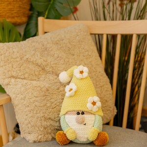 May include: A handmade crochet gnome doll with a yellow hat decorated with white flower accents. The gnome has a light green body, a beige nose, and orange feet. It sits on a grey chair cushion, with a beige pillow in the background.