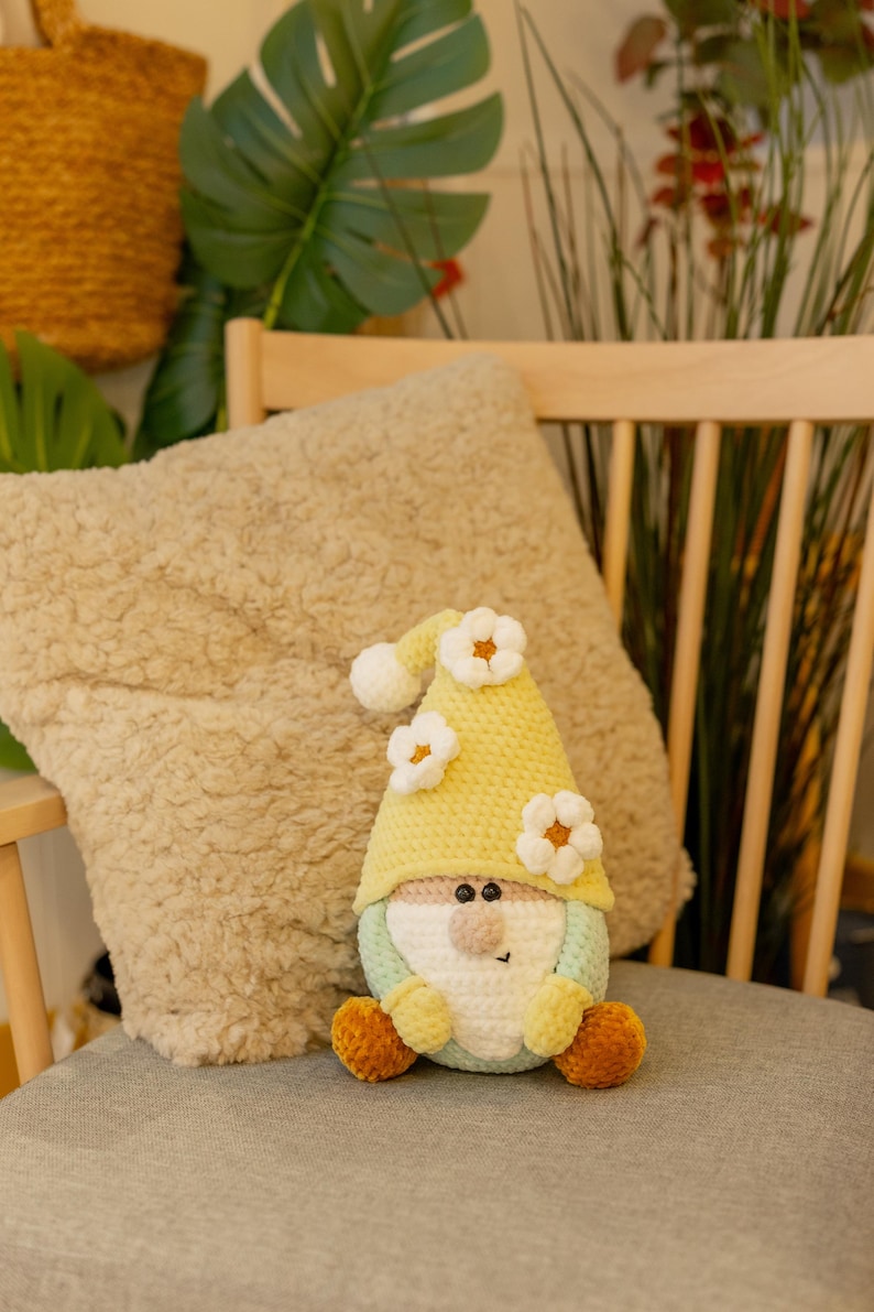 May include: A handmade crochet gnome doll with a yellow hat decorated with white flower accents. The gnome has a light green body, a beige nose, and orange feet. It sits on a grey chair cushion, with a beige pillow in the background.