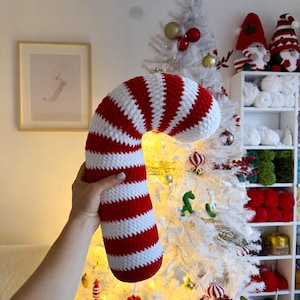 Puede incluir: Una almohada de caramelo de crochet en rayas rojas y blancas. La almohada se sostiene frente a un árbol de Navidad blanco decorado con luces y adornos.