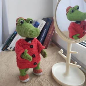 Puede incluir: Un cocodrilo de crochet verde que lleva una chaqueta roja con botones negros y una pajarita blanca. El cocodrilo est&aacute; de pie sobre una superficie marr&oacute;n frente a un espejo blanco.