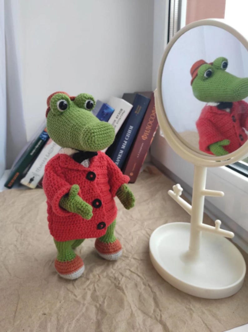 Puede incluir: Un cocodrilo de crochet verde que lleva una chaqueta roja con botones negros y una pajarita blanca. El cocodrilo est&aacute; de pie sobre una superficie marr&oacute;n frente a un espejo blanco.