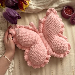 May include: A light pink, handmade crochet butterfly cushion with a ruffled edge design. The butterfly has detailed wings and antennae. The cushion rests on a cream blanket, with purple and yellow tulips in the background.