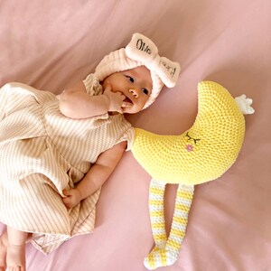 May include: A baby wearing a pink bow headband with the name "Olivia" and a striped dress lies next to a yellow crocheted moon toy with striped legs. The baby is on a pink sheet.