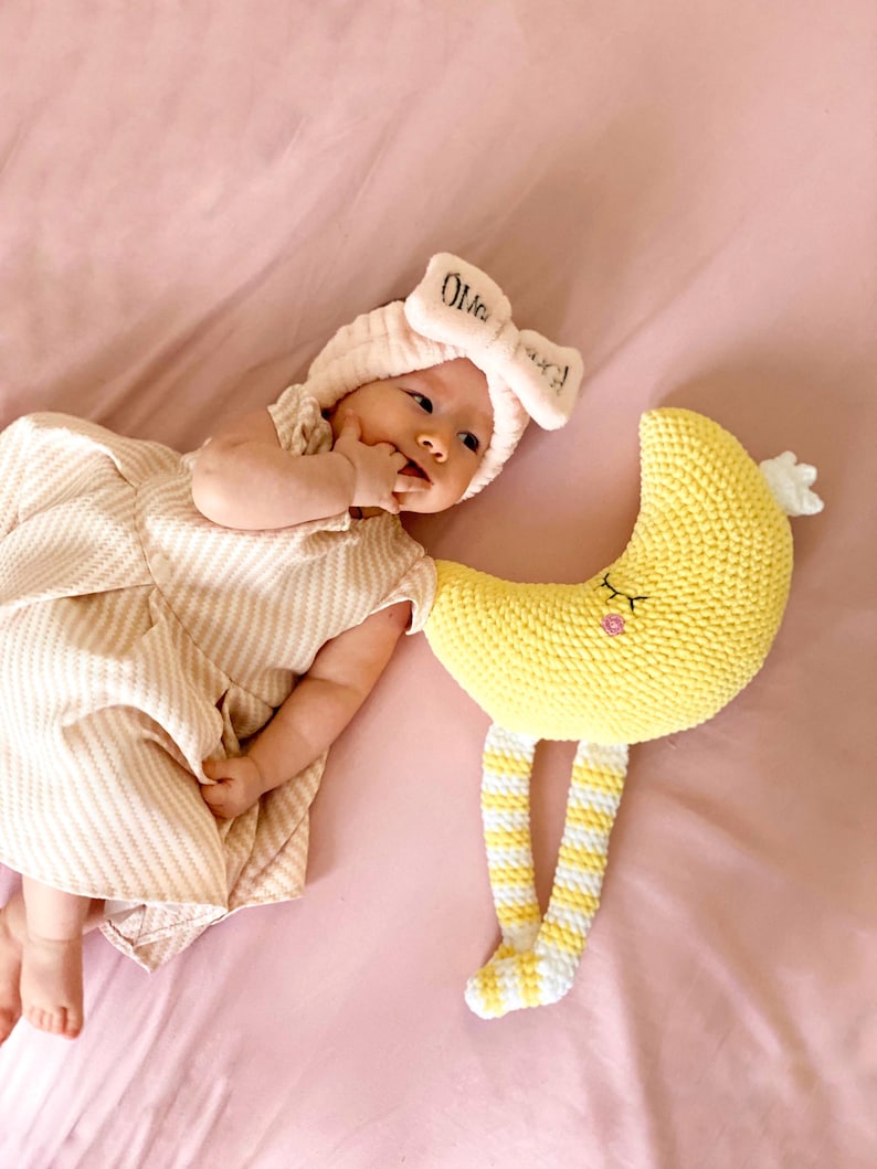 May include: A baby wearing a pink bow headband with the name "Olivia" and a striped dress lies next to a yellow crocheted moon toy with striped legs. The baby is on a pink sheet.