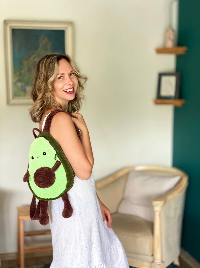 May include: A woman wearing a white dress is carrying a green and brown crocheted avocado backpack. The backpack has a smiling face and brown arms and legs.
