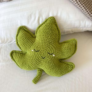 May include: A green, crocheted leaf-shaped cushion with a smiling face and closed eyes. The cushion is made of textured yarn and rests on a white bed with a waffle-weave blanket.