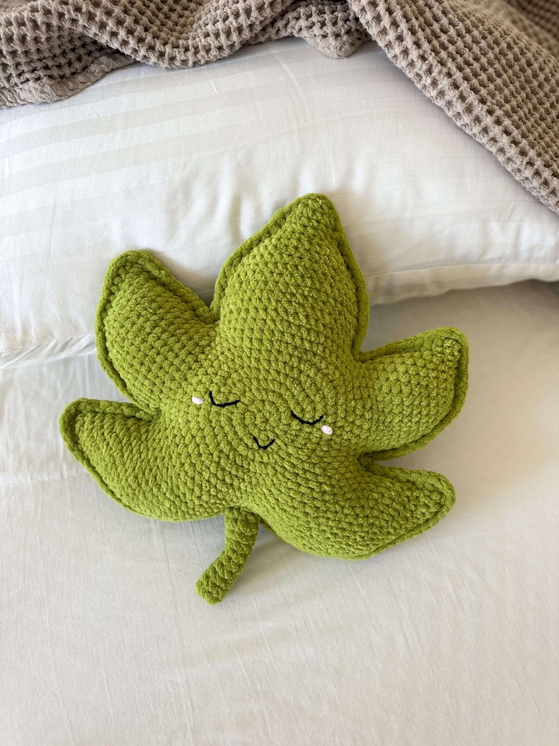 May include: A green, crocheted leaf-shaped cushion with a smiling face and closed eyes. The cushion is made of textured yarn and rests on a white bed with a waffle-weave blanket.