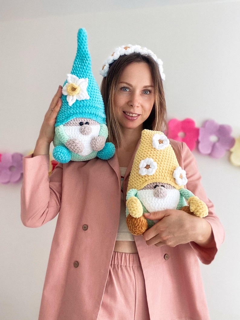May include: Two handmade crochet gnomes. One gnome has a turquoise hat with a white and yellow flower, the other has a yellow hat with white flowers. Both gnomes have white beards, and are held by a person wearing a pink blazer.