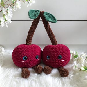 May include: Two plush red cherries with brown stems and green leaves. The cherries have smiling faces with blue eyes and pink cheeks. They are sitting on a white fluffy surface.