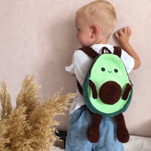 May include: A child wearing a white shirt and blue denim shorts is wearing a green and brown crocheted avocado backpack with a smiling face.