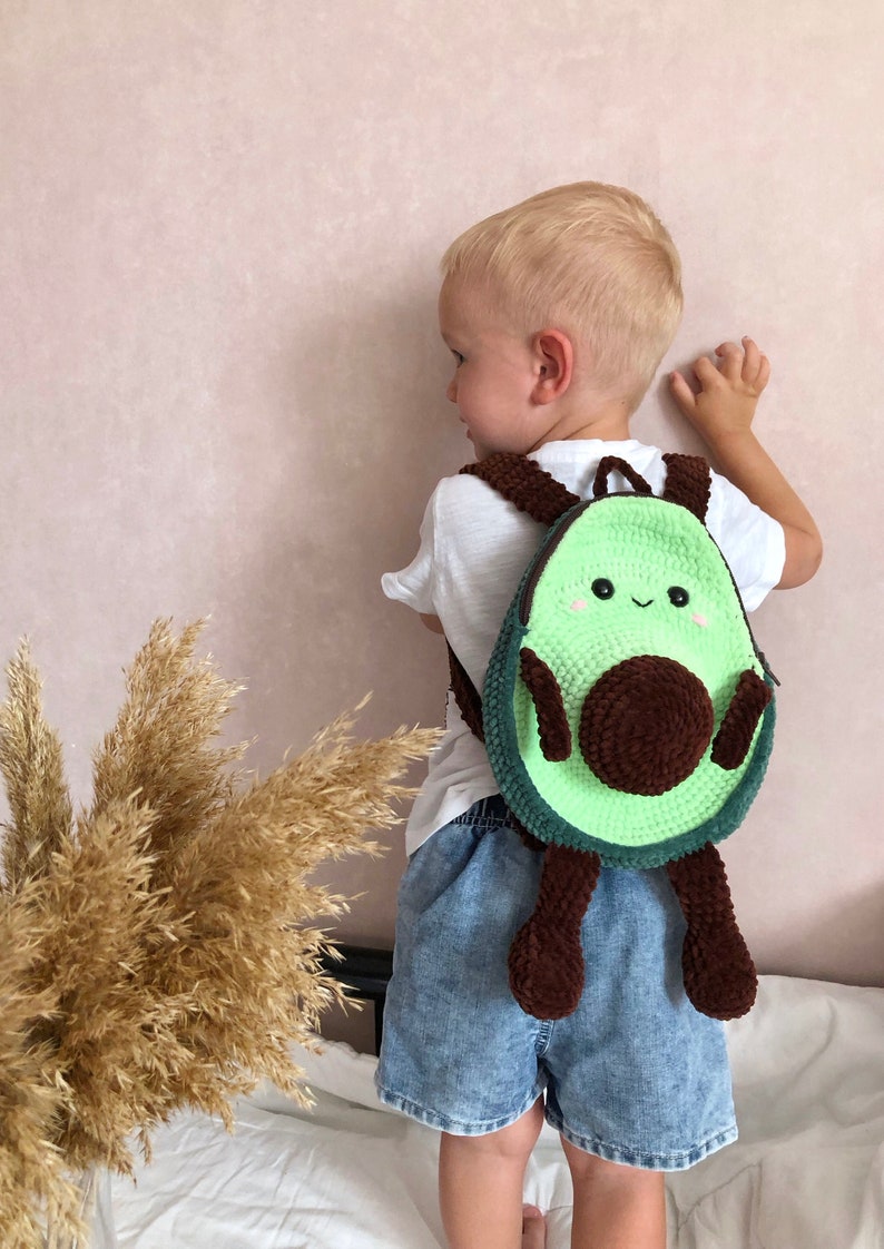 May include: A child wearing a white shirt and blue denim shorts is wearing a green and brown crocheted avocado backpack with a smiling face.