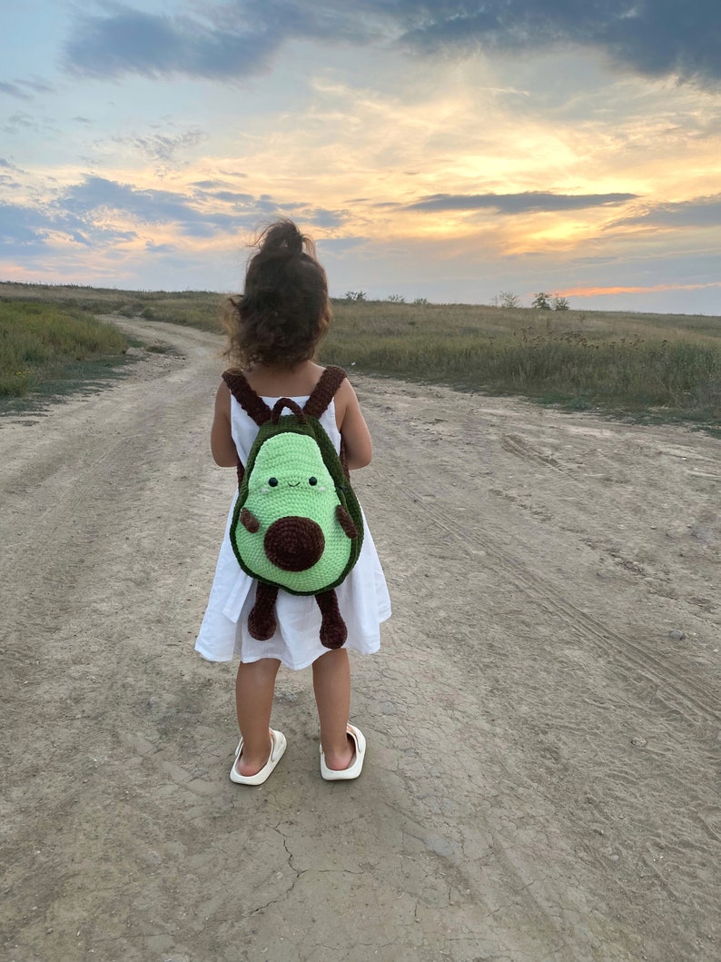 May include: A child wearing a white dress walks on a dirt path with a green and brown avocado-shaped crocheted backpack.