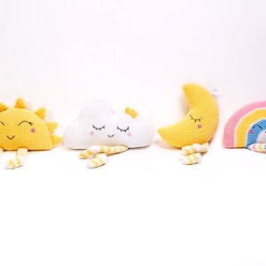 May include: Four crocheted stuffed toys: a yellow sun, a white cloud, a yellow crescent moon, and a rainbow with a blue face. Each toy has a smiling face with closed eyes and yellow and white striped legs.