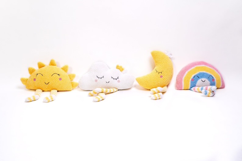 May include: Four crocheted stuffed toys: a yellow sun, a white cloud, a yellow crescent moon, and a rainbow with a blue face. Each toy has a smiling face with closed eyes and yellow and white striped legs.