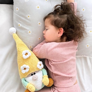 May include: A child asleep in pink ribbed pyjamas, lying on a pillow patterned with daisies. Beside the child is a handmade crochet gnome doll, yellow with white flowers, a white beard, and orange hands.