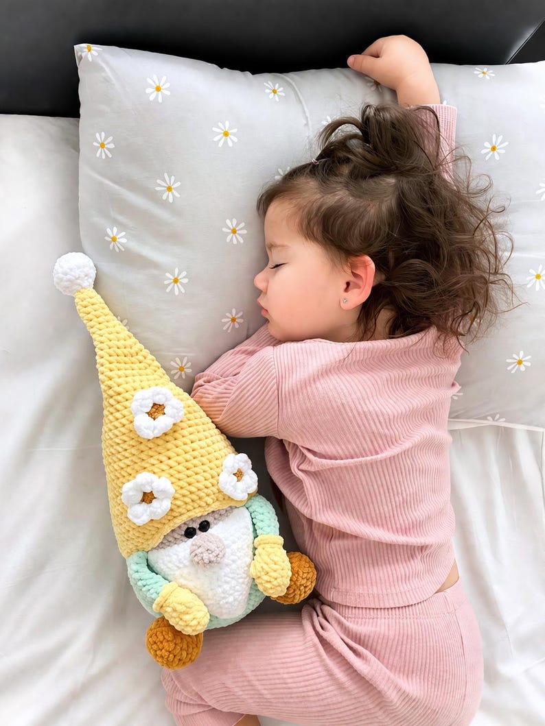 May include: A child asleep in pink ribbed pyjamas, lying on a pillow patterned with daisies. Beside the child is a handmade crochet gnome doll, yellow with white flowers, a white beard, and orange hands.