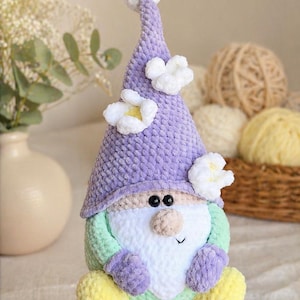 May include: A handmade crochet gnome doll with a lavender hat decorated with white flowers and a white pom-pom. The gnome has a white beard, a green body, and yellow feet. The doll is approximately 20 cm tall.