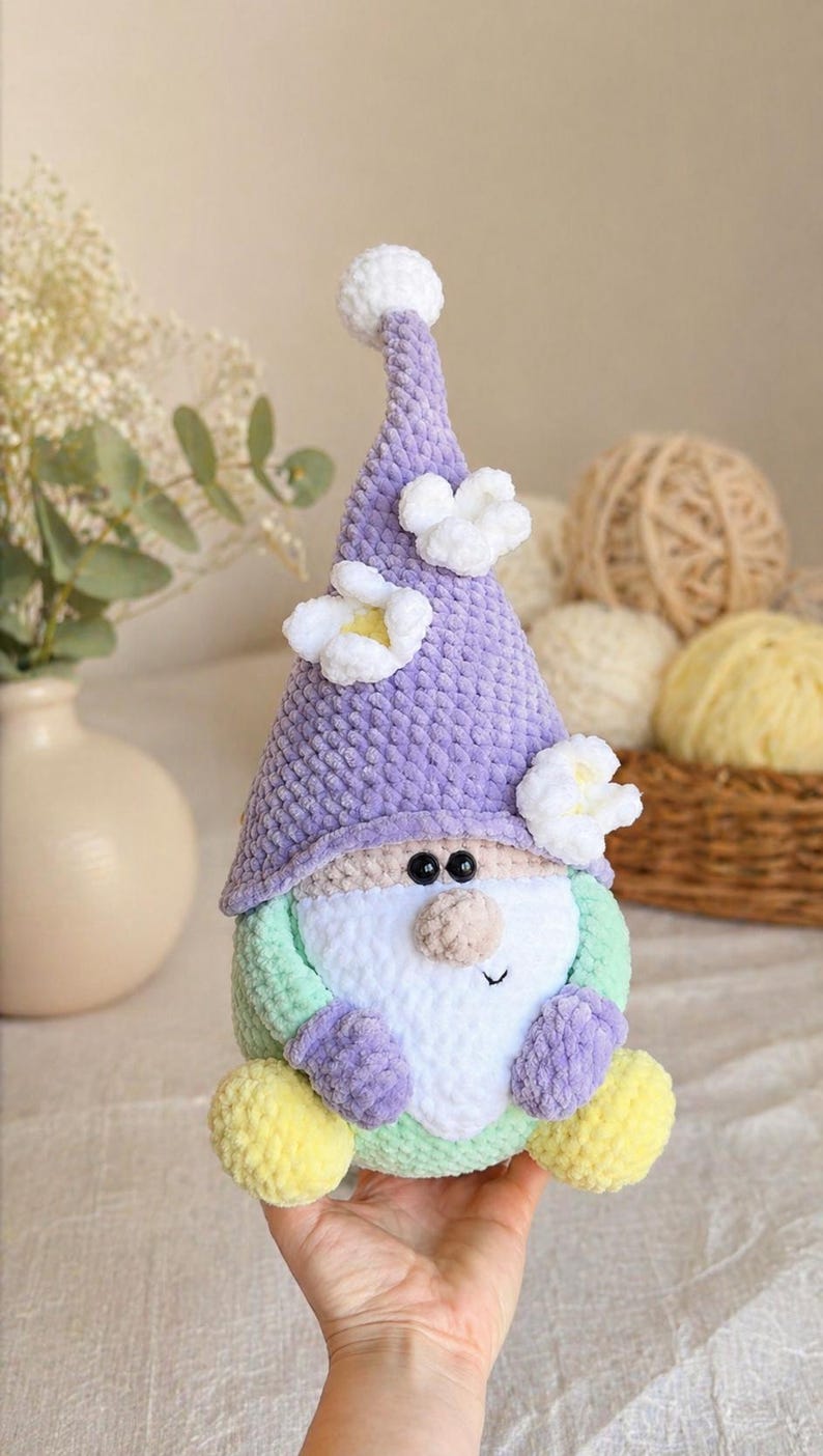 May include: A handmade crochet gnome doll with a lavender hat decorated with white flowers and a white pom-pom. The gnome has a white beard, a green body, and yellow feet. The doll is approximately 20 cm tall.