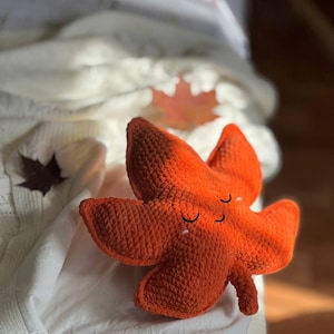 May include: A handcrafted, orange crochet leaf with a smiling face. The plush leaf has a textured surface and is resting on a white surface. The leaf is a fall-themed decorative item.
