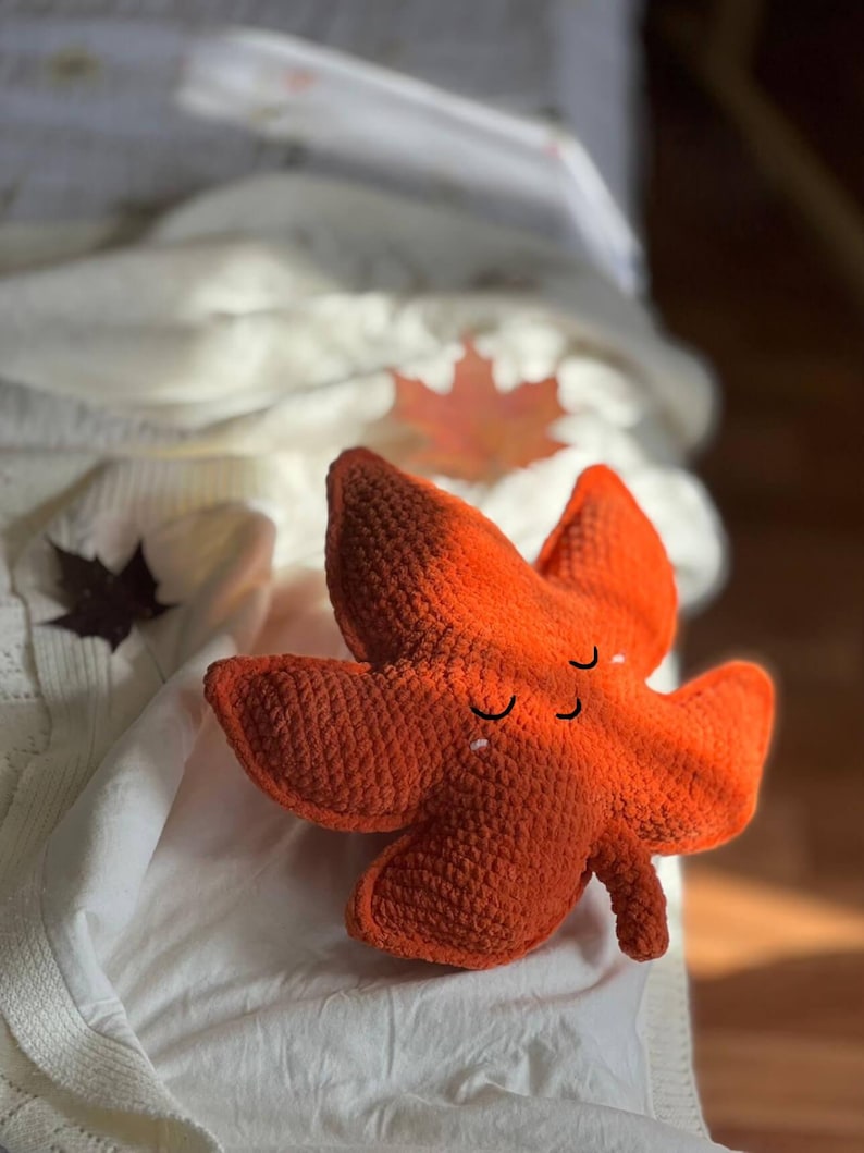 May include: A handcrafted, orange crochet leaf with a smiling face. The plush leaf has a textured surface and is resting on a white surface. The leaf is a fall-themed decorative item.
