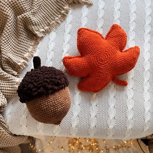 Puede incluir: Una bellota de ganchillo con un sombrero texturizado de color marrón oscuro y un cuerpo marrón claro, junto a una hoja de arce de ganchillo de color naranja brillante. Ambos objetos presentan un patrón de puntos visible y aspecto mullido.