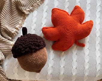 May include: A crochet acorn with a dark brown textured cap and a light brown body, paired with a bright orange crochet maple leaf. Both items are crafted with a visible yarn stitch pattern, creating a soft, plush appearance for these decorative pieces.
