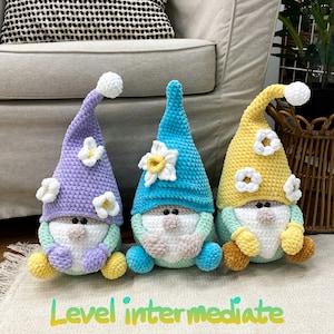 May include: Three handmade crochet gnomes in pastel shades. Each gnome features a pointed hat with a white bobble and a flower detail. The gnomes are positioned on a cream-coloured rug. Text on the rug reads "Level intermediate."