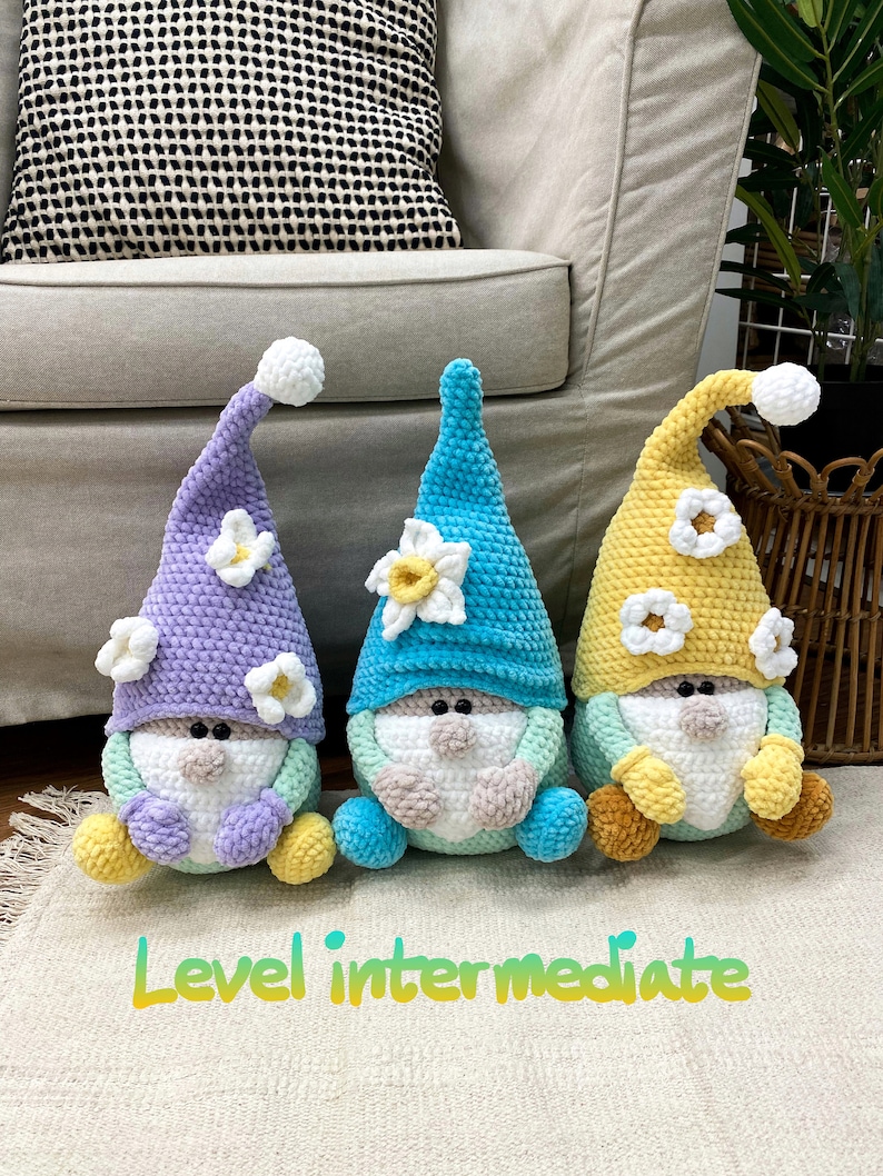 May include: Three handmade crochet gnomes in pastel shades. Each gnome features a pointed hat with a white bobble and a flower detail. The gnomes are positioned on a cream-coloured rug. Text on the rug reads "Level intermediate."