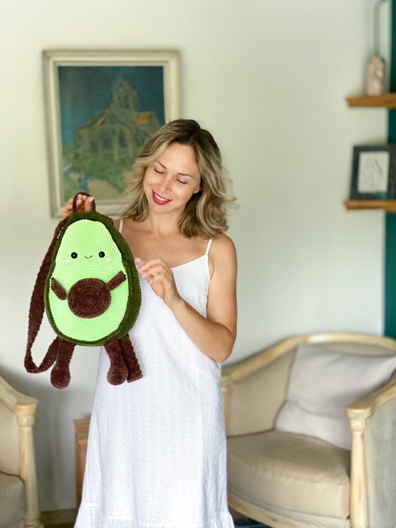 May include: A green and brown crocheted avocado-shaped backpack with a smiling face and a brown pit. The backpack has a brown strap and fuzzy brown legs.