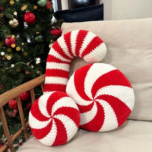 Puede incluir: Un conjunto de tres cojines navideños a rayas rojas y blancas. El conjunto incluye dos cojines redondos y un cojín con forma de bastón de caramelo. Los cojines están hechos de un material suave y texturizado, colocados en un sofá neutro.