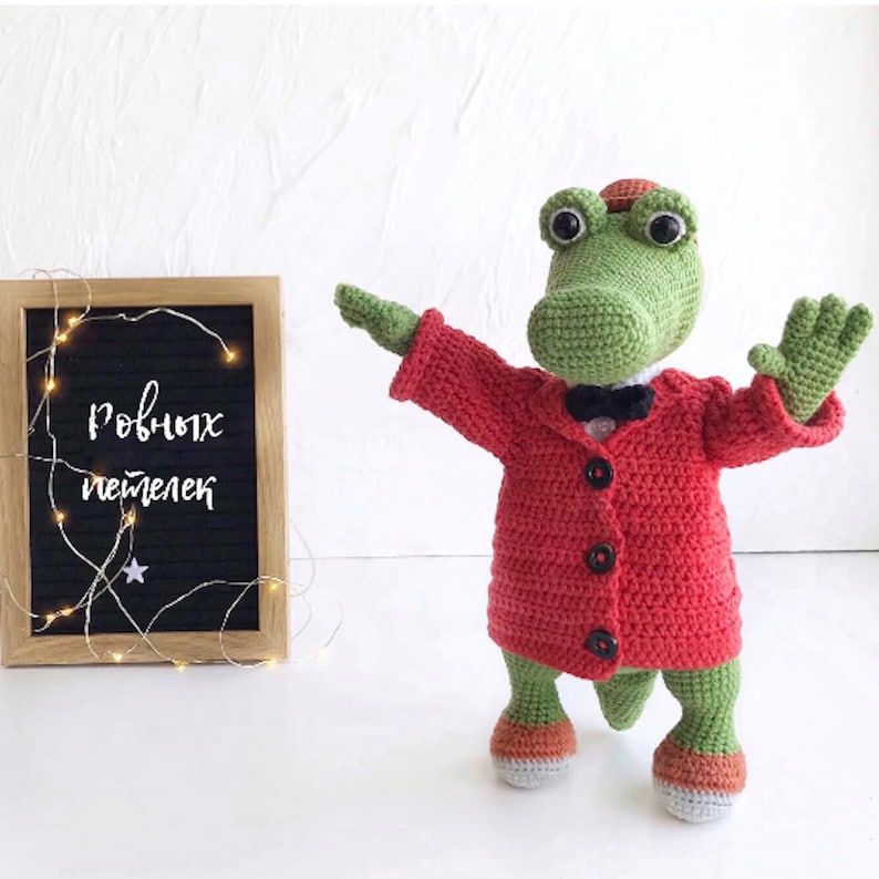 Puede incluir: Un cocodrilo de crochet verde que lleva un abrigo rojo con botones negros y una pajarita negra. El cocodrilo est&aacute; de pie sobre una superficie blanca con un cartel que dice "ро́вных пе́тель" en ruso.