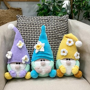 May include: Three handmade crochet gnomes with long, pointed hats and white beards. The gnomes have different coloured hats: lavender, blue, and yellow, each adorned with white flower accents. They are seated on a beige chair with a patterned pillow in the background.