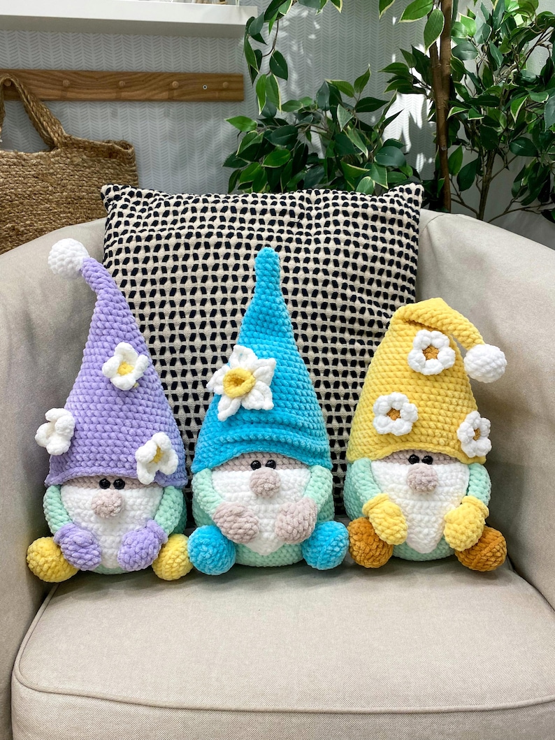 May include: Three handmade crochet gnomes with long, pointed hats and white beards. The gnomes have different coloured hats: lavender, blue, and yellow, each adorned with white flower accents. They are seated on a beige chair with a patterned pillow in the background.
