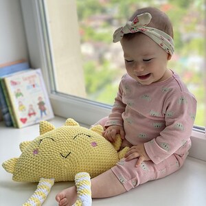May include: A baby wearing a pink outfit with a floral headband is sitting on the floor and holding a yellow crocheted sun toy with white stripes.