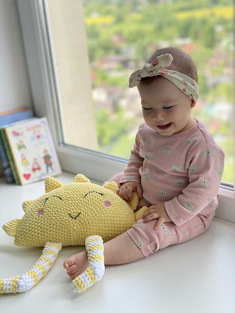 May include: A baby wearing a pink outfit with a floral headband is sitting on the floor and holding a yellow crocheted sun toy with white stripes.