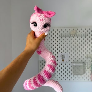 Könnte beinhalten: Ein rosa gehäkeltes Schlangenspielzeug mit einer rosa Schleife auf dem Kopf und großen schwarzen Augen. Die Schlange ist in einer Hand zusammengerollt.