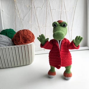Puede incluir: Un cocodrilo de crochet verde que lleva una chaqueta de crochet roja. El cocodrilo est&aacute; de pie sobre una superficie blanca con una cesta de hilo en el fondo.