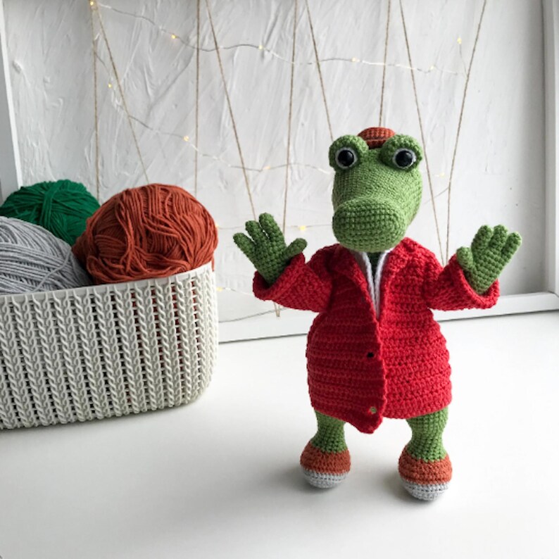 Puede incluir: Un cocodrilo de crochet verde que lleva una chaqueta de crochet roja. El cocodrilo est&aacute; de pie sobre una superficie blanca con una cesta de hilo en el fondo.