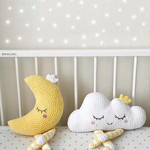 May include: Crocheted nursery decor featuring a yellow crescent moon and a white cloud, both with closed eyes and small floral accents. The moon and cloud have striped yellow and white legs. The items are placed on a crib.